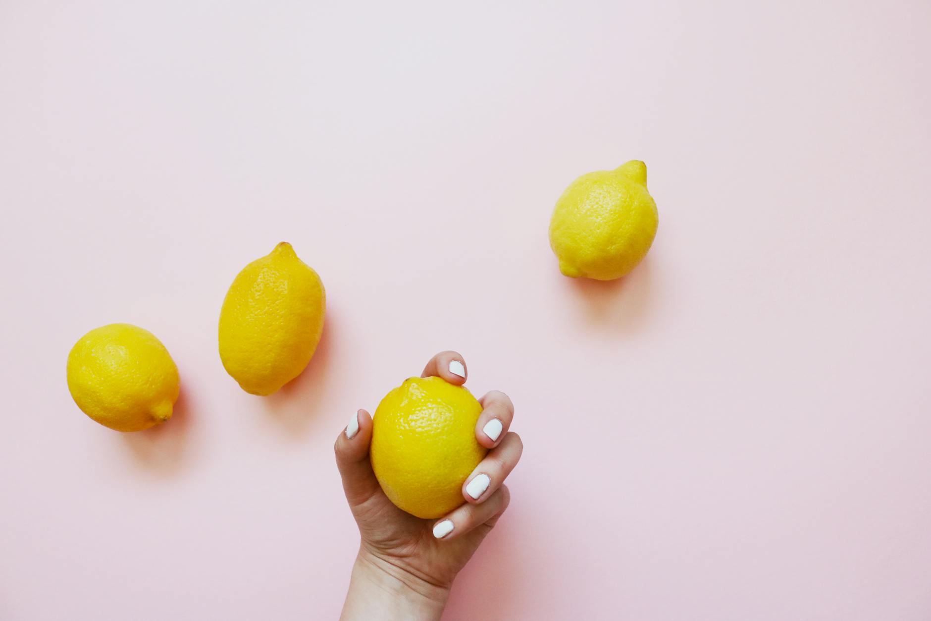 A hand holding a bright yellow lemon on a soft pink background, surrounded by additional lemons
