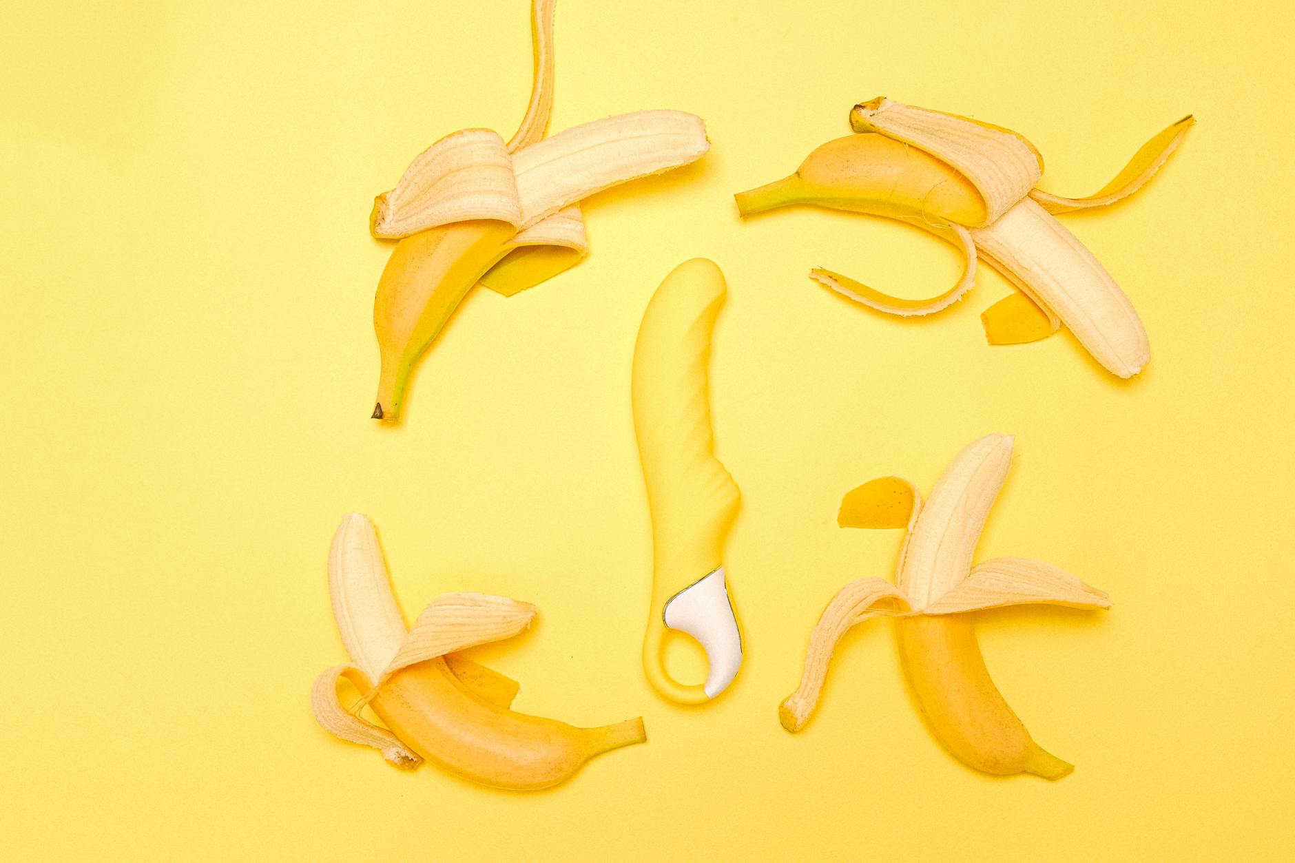 Creative flat lay of a yellow silicone vibrator surrounded by peeled bananas on a yellow background.